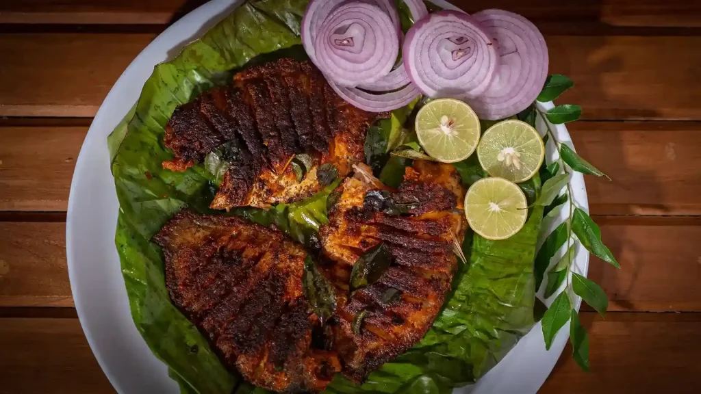 Spicy Kerala-style fish fry served on a banana leaf with sliced red onions, fresh lime halves, and curry leaves on a white plate placed on a wooden table.
